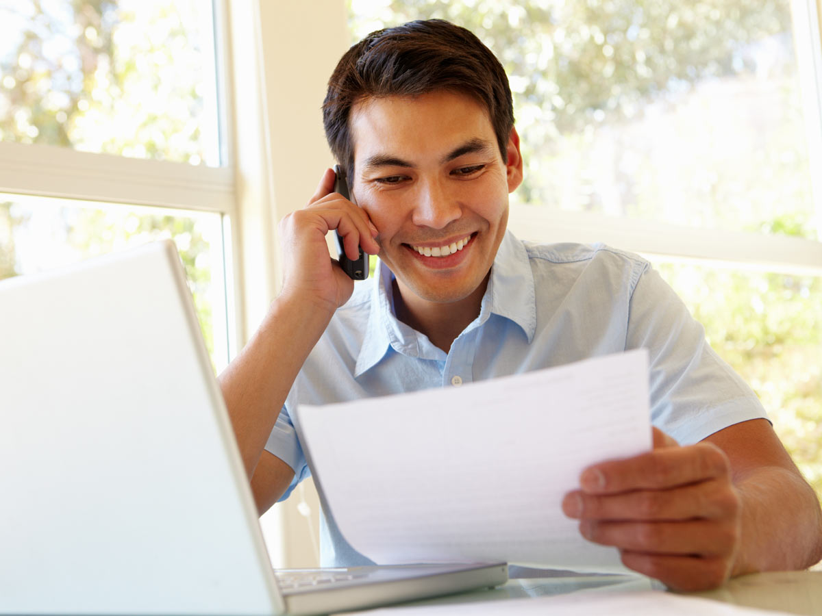 A young man gets an insurance quote by phone. He sits with his laptop open in front of him and looks over his insurance policy.